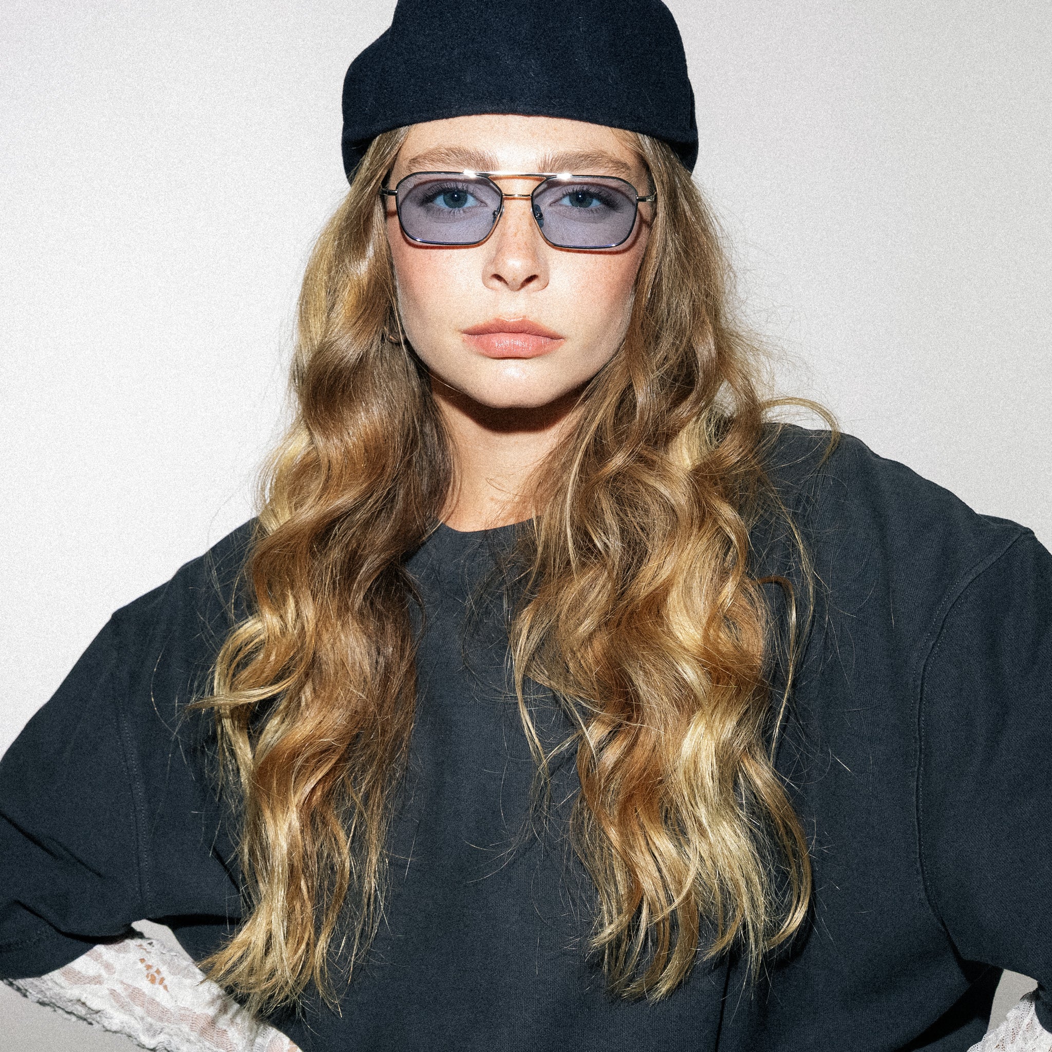 Person with long wavy hair wearing a black beanie and sunglasses against a plain background