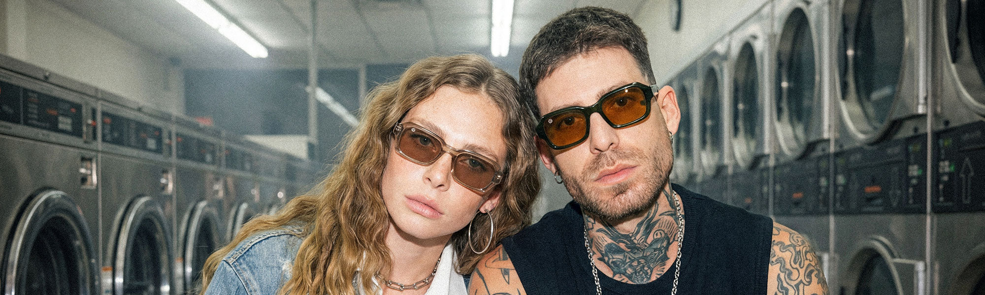Two models wearing light-tinted sunglasses in a laundromat, close-up portrait with washing machines in the background
