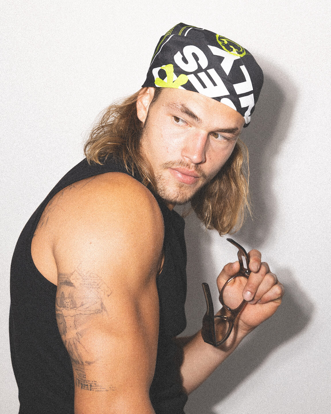 Man with long hair wearing a bandana and holding sunglasses against a white background